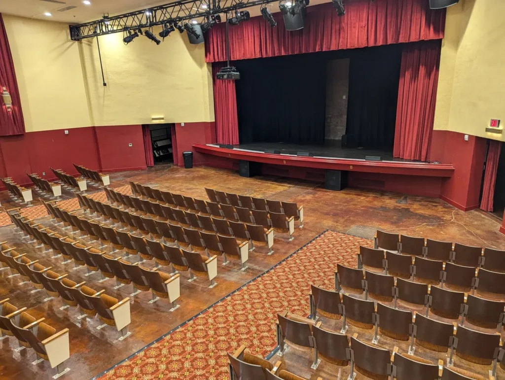 The Lyric's auditorium from the balcony: rows of wooden seats either side of a patterned carpet aisle, facing the stage and its red curtains