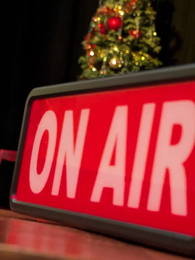 A red ON AIR sign beside a Christmas tree on stage