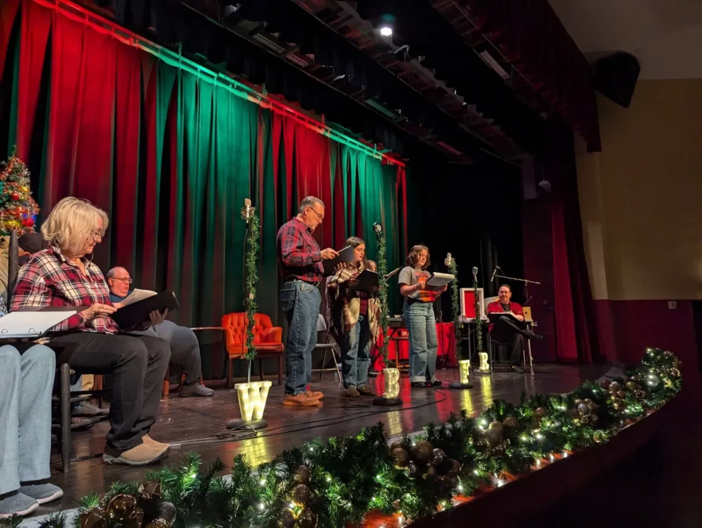 Performers reading at microphones on stage, with lit greenery along the front of the stage